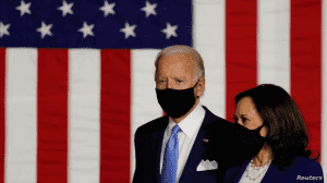 An image of Joe Biden and Kamala Harris standing next to one another. They wear masks and stand in front of a large American Flag. The text 