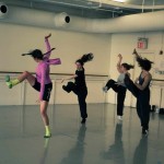 In a dance studio, dancers are lift one leg bending, and flexing it, letting their arms and hair fly free.