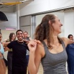 A picture of a dance class. There is a woman in front smiling with her arm around the shoulders of her dance partner.