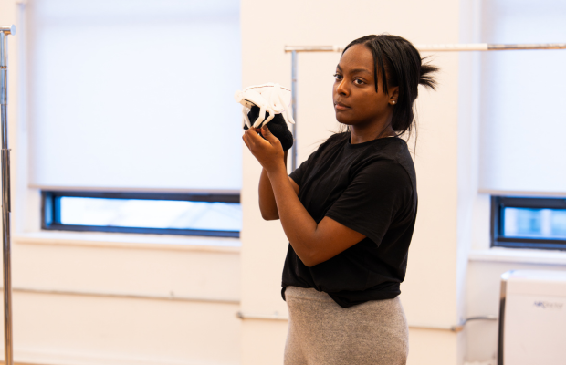 A dancer in a black shirt and gray pants holds a cricket puppet in a rehearsal room.