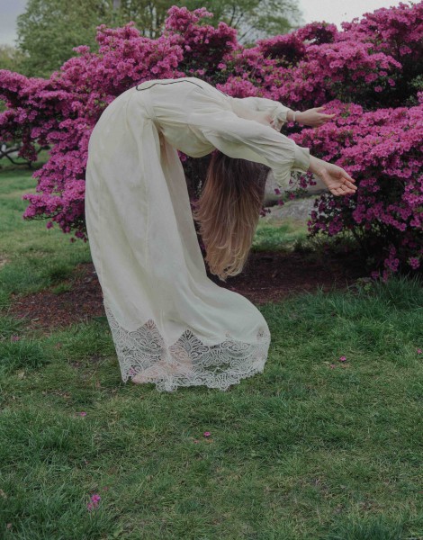 Woman in a white long dress lean back in a dance in front of pink blooming bush behind 