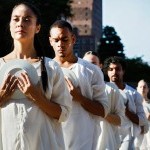 Dancers in white clasping a white ceramic plate to their chest in line formation on outdoor plaza
