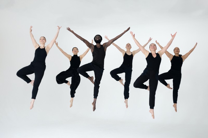 Six dancers dressed in black dancewear jumping in the air with arms extended and joyful expressions.