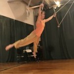 Woman with arms and legs out stretched while flying by hanging on to two trapeze bars, wearing orange shirt and flowing pants