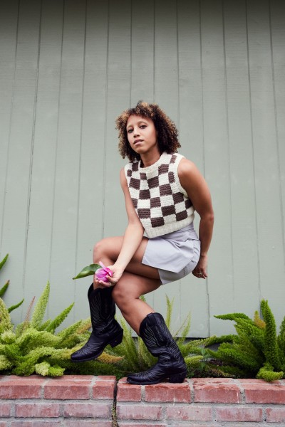 A brown, curly haired woman wearing a patterned blouse and cowboy boots folds her right arm over her left leg while twisting.