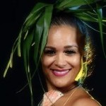 Native Hawaiian/Chamorro with red lipstick on smiling, hair is in a high bun with a green leaf fringe attached and wearing a she