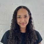 Aryanna Allen, a person with medium brown skin and long dark curly hair, stands in front of a white brick wall. 