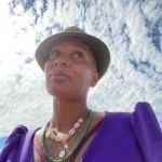A honey colored, bald black woman wearing an evergreen colored hat in a purple jumpsuit with a cowry shell and beaded necklace