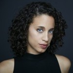 A woman with dark brown, curly hair and blue eyes looks at the camera