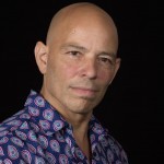 A man with a bald head wearing a blue paisley-type print shirt looks at the camera