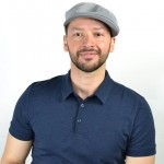 A man wearing a dark blue polo shirt and light gray cap smiles softly at the camera