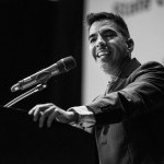Older male-presenting person wearing a dark suit and a white beard stands smiling at a podium. 