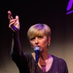 upper body shot of Lucy Sexton at mic, hand raised in speech, blonde hair, grey top