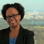 Light skinned, black woman with dark curly hair. Wearing a black blazer, a blue blouse, and eyeglasses. Standing with crossed ar