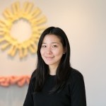 Headshot of Michelle Bae in a black sweater smiling in front of John Ahearn’s yellow and orange plaster cast sculptures.