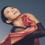 Muna Tseng portrait with her head tilted back and her arms arm raised with her hand curled with an orange scarf.