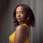 Profile to the camera is a brown-skinned Black woman with deep red shoulder length hair in a mustard yellow sleeveless dress and