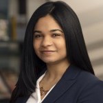 Patricia, with long dark hair, wears a white V-neck and a jacket, and smiles slightly for the camera.