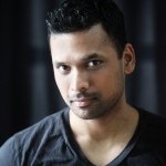 Brown skinned, Indian American, wearing a black V-neck tee with short hair combed neatly up. Angled perspective of the face