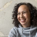 Happy smiling cis-woman with dark curly hair, light-skin, and of mixed heritage.
