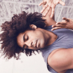 Joey is rockin’ brown skin and an afro with black hair. He is back bending with his palms splayed to frame his face.