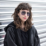 A white woman with a curly brown shag haircut, black leather jacket, and slightly tinted aviator glasses in front of an industri
