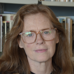 Red-haired white woman, wearing a black sweater, standing against a shelf of books.