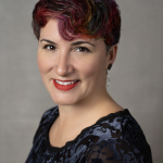 White queer femme with multicolored short hair, smiling at the camera, wearing a red lip with a velvet top with a lace design.