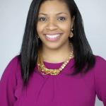 Kellee, a non-disabled butterscotch-skinned woman with long dark brown hair, is facing the camera, smiling in front of a gray ba