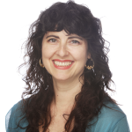 Maya has dark curly hear and wears glasses, looking to the camera.