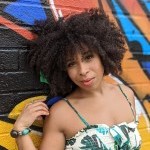 Standing against a colorful graffiti mural, Shari is smiling softly with a slight tilt of her head towards the camera