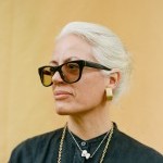 Yanira is a light-skinned Latine person wearing black thick rimmed glasses and her white hair pulled back. Yellow background.