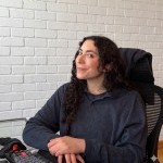 A portrait photograph of a smiling person with light skin and long, dark curly hair, sitting in an office chair at a desk with a