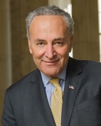 A headshot for Senator Chuck Schumer. Chuck looks towards the camera wearing a grey blazer with an American flag pin on it, a blue shirt and a yellow tie with blue spots on it.