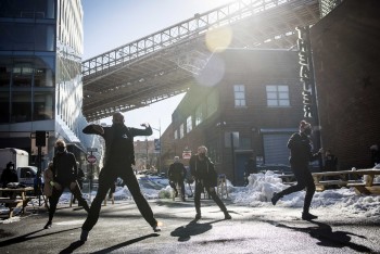 An intersection in Brooklyn. The underside of the manhattan bridge can be seen in the background. A group of dancers pictured in motion wearing winter gear and face coverings. 