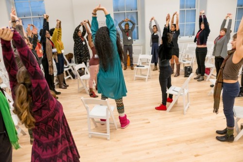A large group of people, standing in a circle in a dance studio, stretch their arms upward.