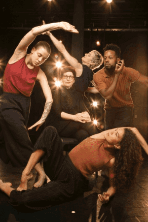 Five dancers surround a woman sitting in a chair in a studio lit as if for performance