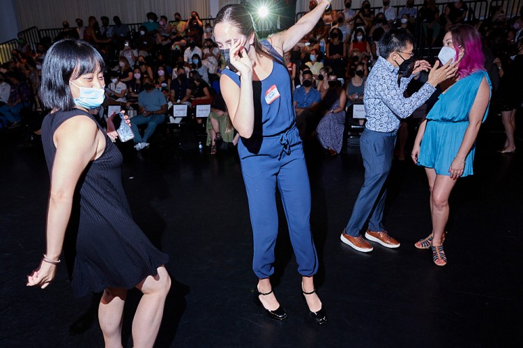 A woman in a blue navy suit dances front and center. Other attendees are seen dancing and talking to each other at the Dance/NYC DWR Initiative Launch Event.