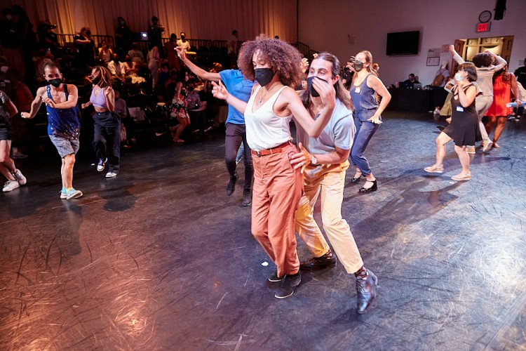 Two attendees are captured dancing with each other to the music featured at the Dance/NYC DWR Initiative Launch Event. Behind them, many other attendees are seen dancing in the photo.
