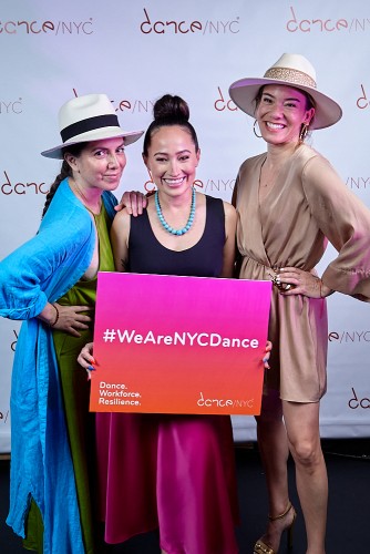 Three women joyously take a picture together during the Dance/NYC DWR Initiative Launch Event, including Dance/NYC’s Executive Director Alejandra Duque Cifuentes who is standing in the middle holding a poster that reads ‘#WeareNYCDance’ front and center. On the bottom right of the poster it reads ‘Dance.Workforce.Resilience’ and the bottom left is the Dance/NYC logo. Behind them is a step-and-repeat with the Dance/NYC logo.