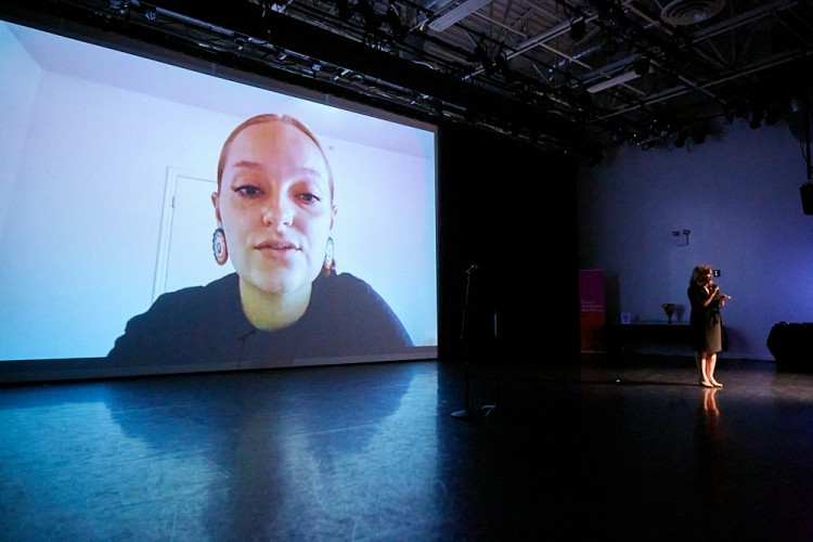 River Whittle, an artist, is captured talking on a big screen at the Dance/NYC DWR Initiative Launch Event. They are wearing a black top and round colorful earrings, and their hair is pulled back.