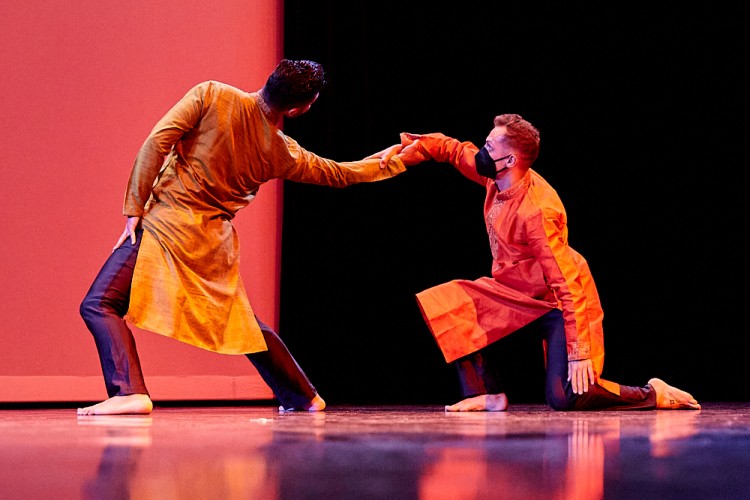 Two members of Rovaco Dance Company are captured performing in front of the audience at the Dance/NYC DWR Initiative Launch Event. They are wearing orange tunics and dark pants, joining hands, one leaning backwards, the other kneeling.