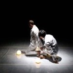 two dancers wearing white costumes kneel next to two rounded white light lamps