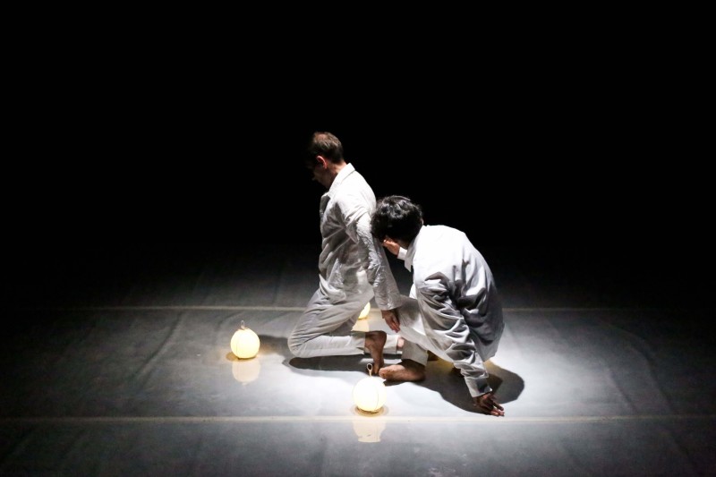 two dancers wearing white costumes kneel next to two rounded white light lamps