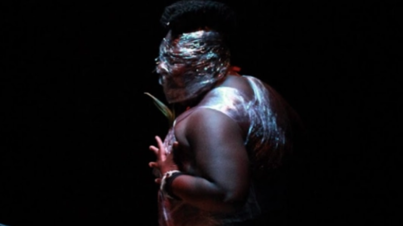 a person wrapped in cellophane looks to the left with their hand against their chest