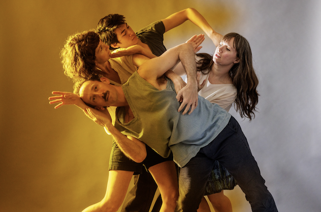 four dancers create angular shapes, their bodies intertwine, a soft yellow light fills the left side of the image 