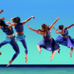 An ensemble of 6 dancers airbound in a spiraling jump in unison onstage.