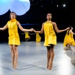 Two dancers in yellow dress jumps solemnly with their arms reaching out to the side. 