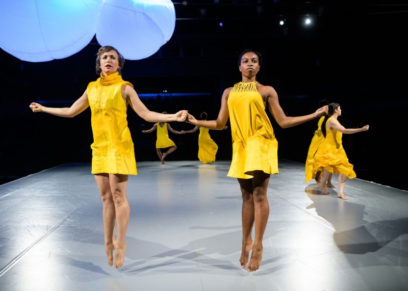 Two dancers in yellow dress jumps solemnly with their arms reaching out to the side. 