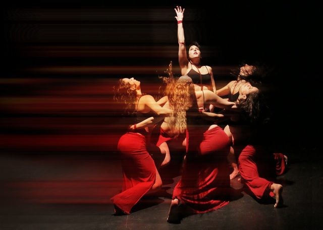 Dancers in black and red costumes form a circle in the dark, at the center of which one dancer stands tall and lifts her hand.
